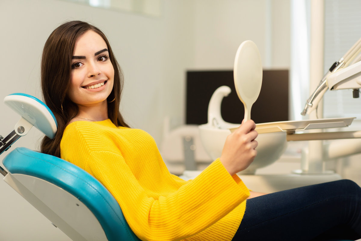 Young happy woman client looking at the mirror with toothy smile at the dental office.